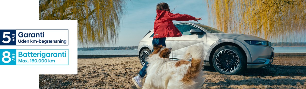 Pige leger med hund på strand foran parkeret Hyundai IONIQ 5 med "Hyundai-garanti" logoer henover