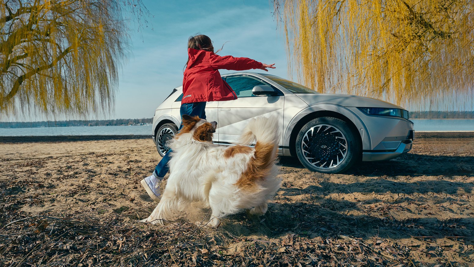 Pige i rød jakke leger med hvid hund på en strand foran parkeret Hyundai IONIQ 5 